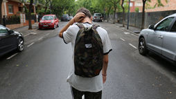 Person with camo backpack walks down a tree-lined street, hand on head. Cars parked on both sides. Overcast day.