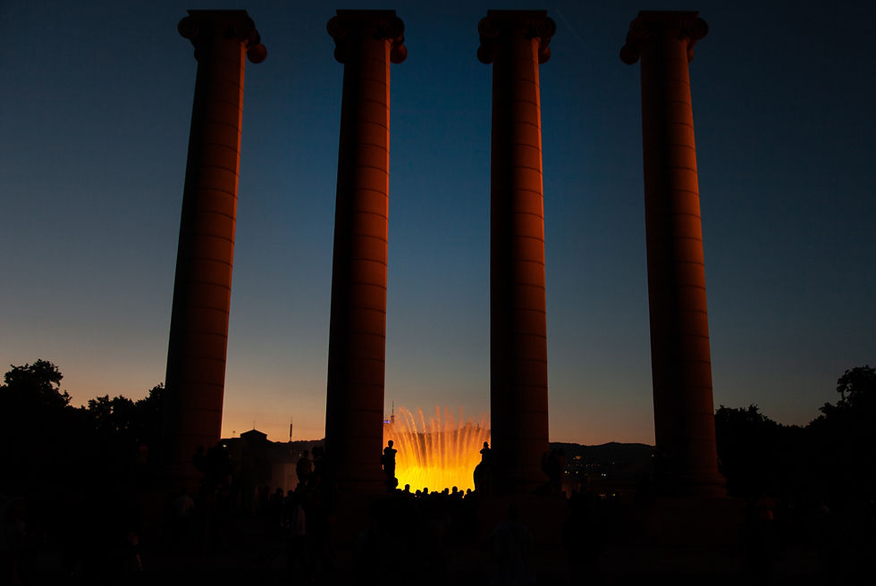 Silhouetted people between four large columns at sunset, with an illuminated fountain glowing orange in the background, creating a serene mood.