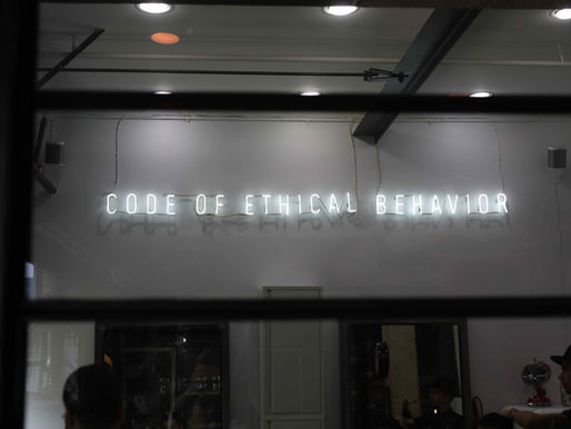 Neon sign reads "CODE OF ETHICAL BEHAVIOR" on a white wall. The setting appears to be indoors with dim lighting and ceiling lights.
