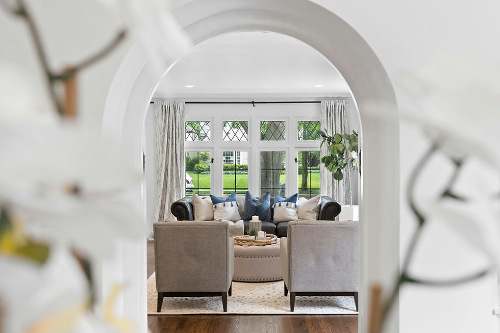 Bright living room viewed through archway, featuring two gray chairs, a dark sofa with blue pillows, and large windows overlooking greenery.