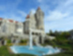 Castle-like building with red roofs under a blue sky. A fountain splashes in the foreground, surrounded by green hedges and gardens.