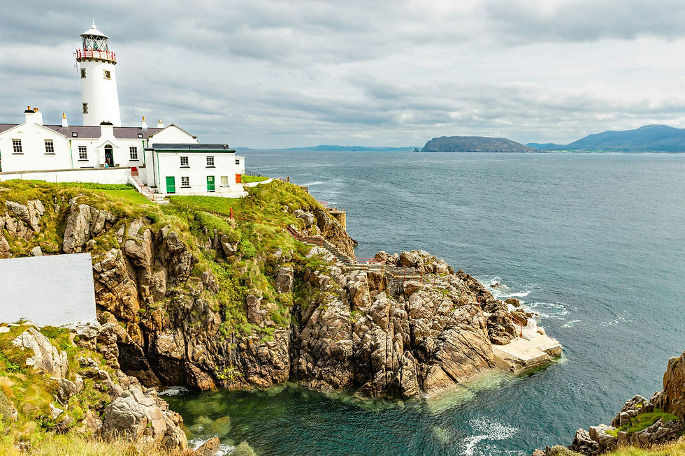 White lighthouse on a rocky cliff above a calm sea, with distant hills and cloudy sky in a quiet coastal scene
