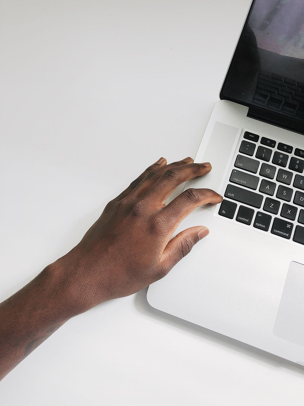 A hand rests on a silver laptop's palm rest, set on a white surface. The keyboard is black with visible keys. The scene is minimalistic.