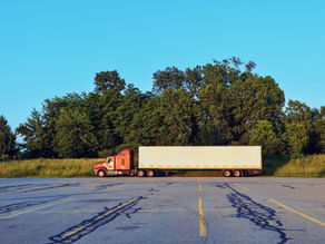 Red semi-truck with white trailer parked on an empty lot, bordered by lush green trees under a clear blue sky.