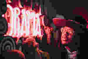 Neon Letters Spelling the Word Drama in Red light. It looks like the sign is on a Fair Ground Wagon side, so you can imagine that this is advertising a Dramatic Adventure insie. A Man wearing Round Glassing and a Wide Brimmed Hat is looking at the sign and you see the red light reflecting on his Glasses and the red glow from the light on his face. Teddy Bears are hanging below and to the side of the Neon Sign Too so making it look quite threatircal