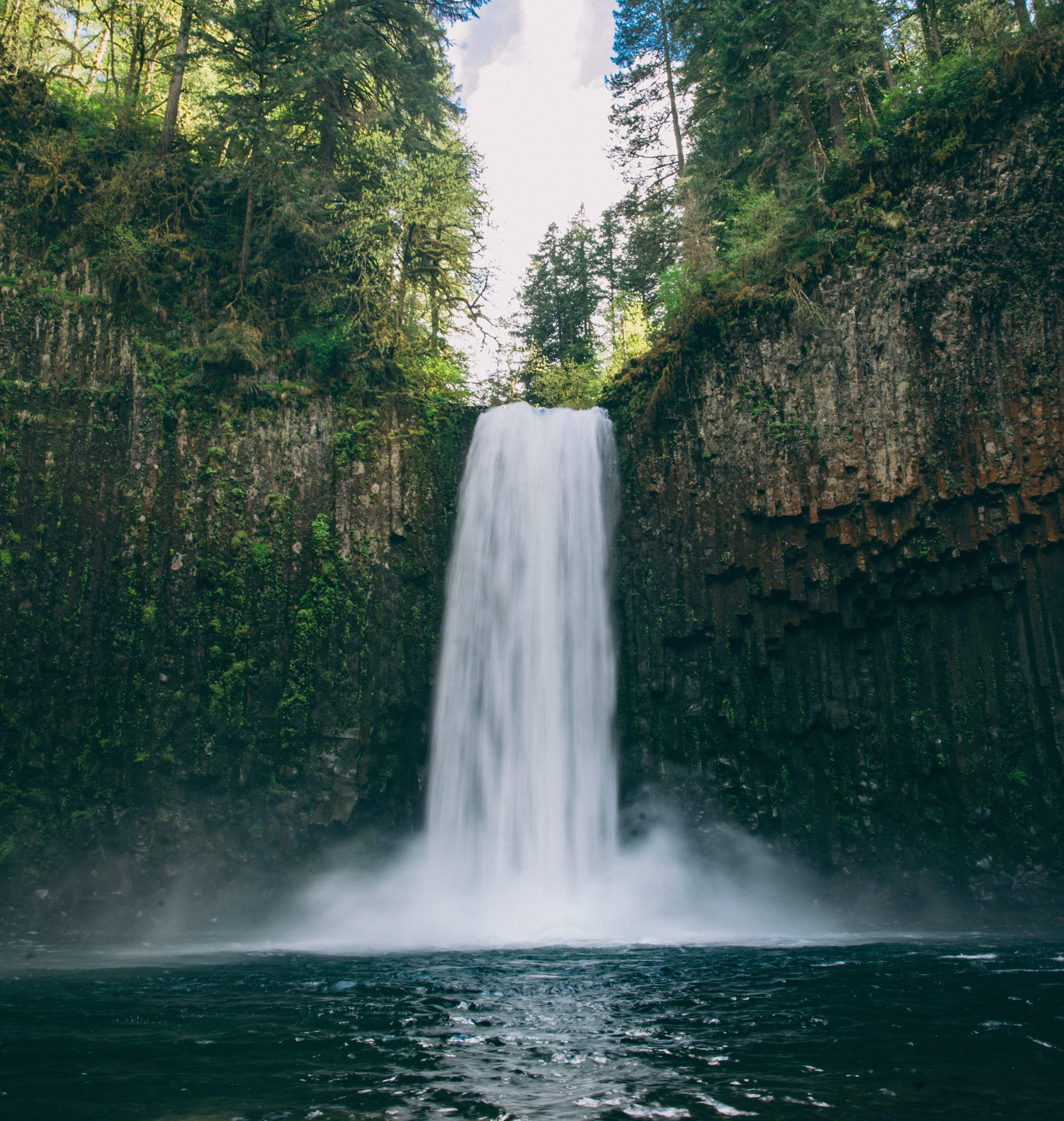 Tall waterfall plunges into a rocky pool in a forest canyon, framed by evergreen trees and mist under a bright blue sky. How the energy and health begins with spirit and flows downward through mind, emotions and physical body.
