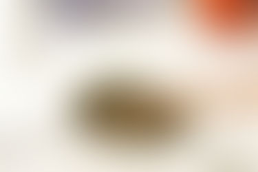 Bowl of mixed dried herbs with a wooden spoon on a light surface. Background shows blurred lavender and a hint of a red drink.