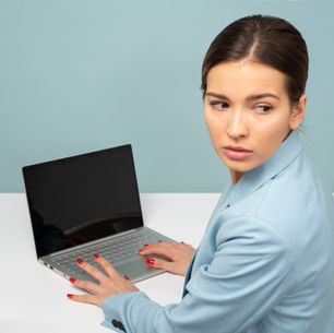 Woman in a light blue blazer looks over her shoulder, sitting at a laptop on a white desk. Background is light blue, mood is curious.