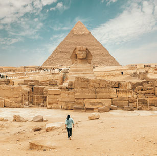 A visitor approaches the Great Sphinx and the Pyramid of Khafre under a bright, cloud-dappled sky in Giza, Egypt.