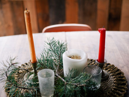 Candles on a woven mat with pine branches. Orange and red taper candles in holders, plus a white candle. Wooden background, cozy mood.