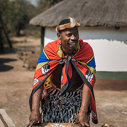 Man in traditional Zulu attire near a thatched hut