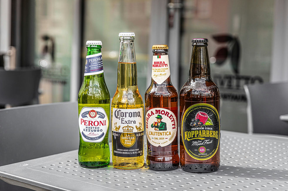 Four beer and cider bottles on a metal table: Peroni, Corona, Birra Moretti, and Kopparberg, with blurred café background.