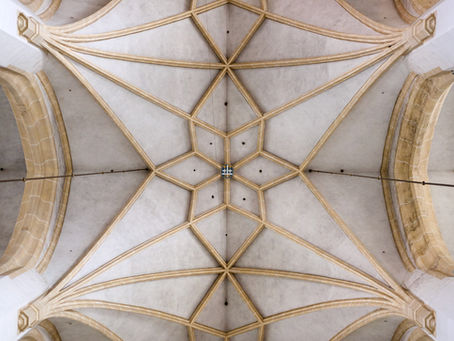 Vaulted ceiling of a church with intricate geometric patterns and a central coat of arms. Beige and white tones dominate the design.