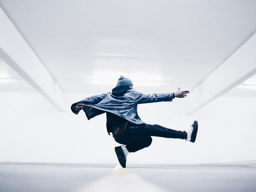 A dancer in a gray beanie and denim jacket leaps midair in a bright white room, arms outstretched.