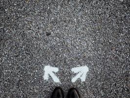 Black shoes on gravel facing two white arrows pointing left and right. The scene suggests a choice or crossroads, evoking contemplation.