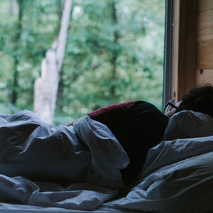 Person asleep under white blankets in a cozy bed beside a large window overlooking a green forest