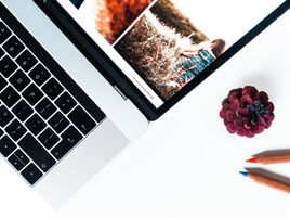 Laptop on white desk showing a photo with find me on Instagram, beside a pinecone and two colored pencils