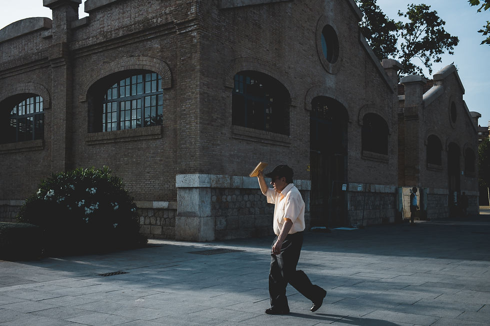 Man walking past a brick building holds a book over his head in sunlight. Trees and another person are visible in the background.