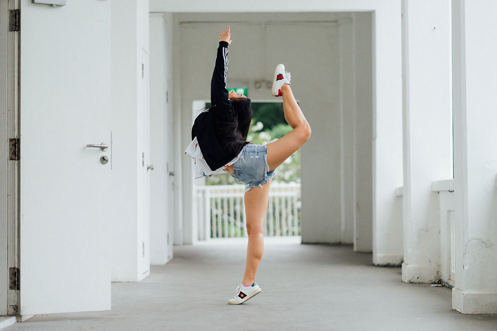 A person performs a high leg stretch in a corridor with white walls, wearing a black jacket, denim shorts, and sneakers. Feels dynamic.