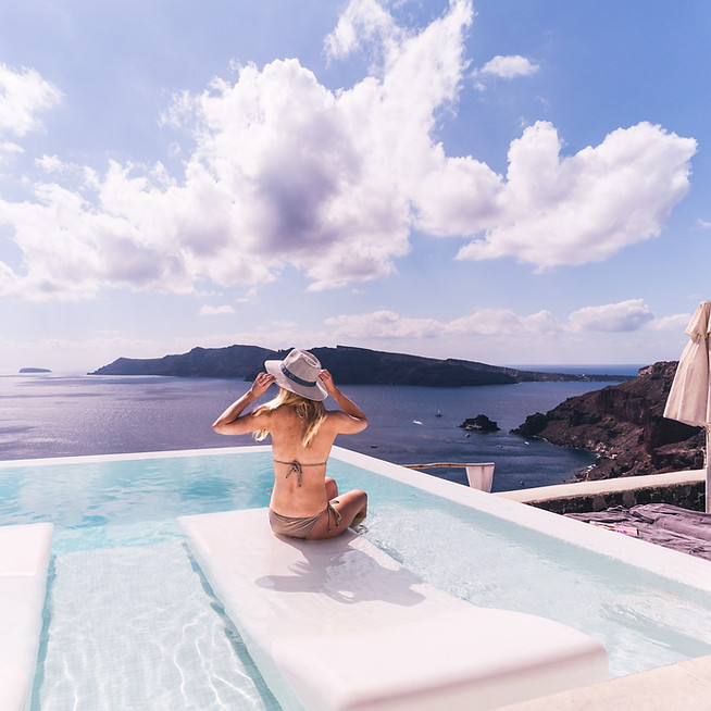 Women in sunhat sitting in pool on white lounger overlooking ocean in Santorini, Greece.
