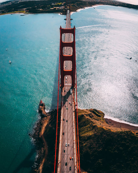 Aerial view of the Golden Gate Bridge with cars crossing. Blue water surrounds, green hills in the background, under a clear sky. Vibrant mood.