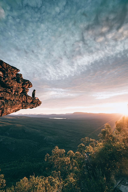 Man sitting on a cliff edge at sunrise, overlooking vast forest and sky, symbolising the soul’s quiet search for purpose beyond the veil
