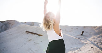 Blonde woman standing in a desert landscape with arms raised, facing away from the camera. 