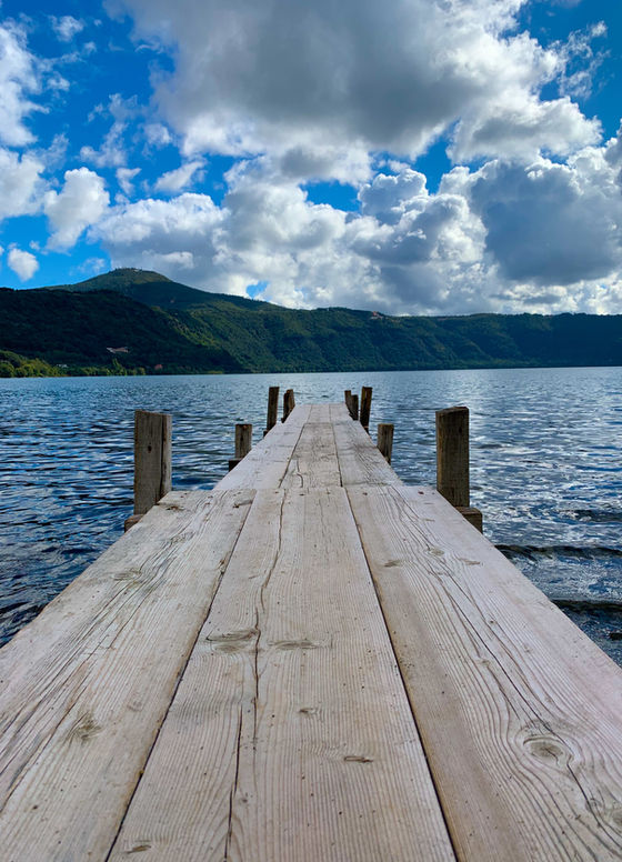Wooden dock extends into a calm lake toward green hills under a bright blue sky with large white clouds.
