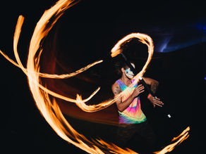 Fire dancer in tie-dye shirt and skull face paint spins flaming staff, orange light trails against dark background.