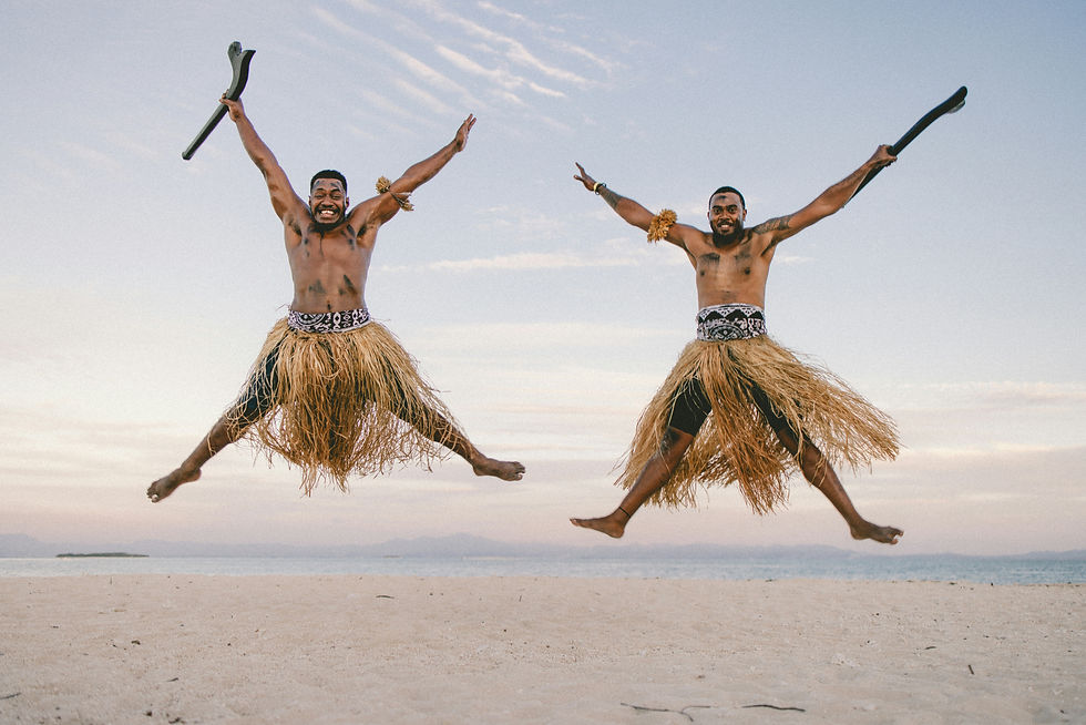 Two men in traditional outfits joyfully jump on a sandy beach, holding wooden clubs against a clear sky.