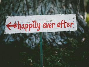 A white wooden sign with red text, "happily ever after," and an arrow stands outdoors against a tree trunk, evoking a cheerful mood.