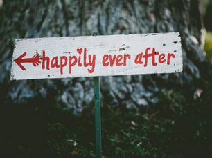 A white wooden sign with red text, "happily ever after," and an arrow stands outdoors against a tree trunk, evoking a cheerful mood.