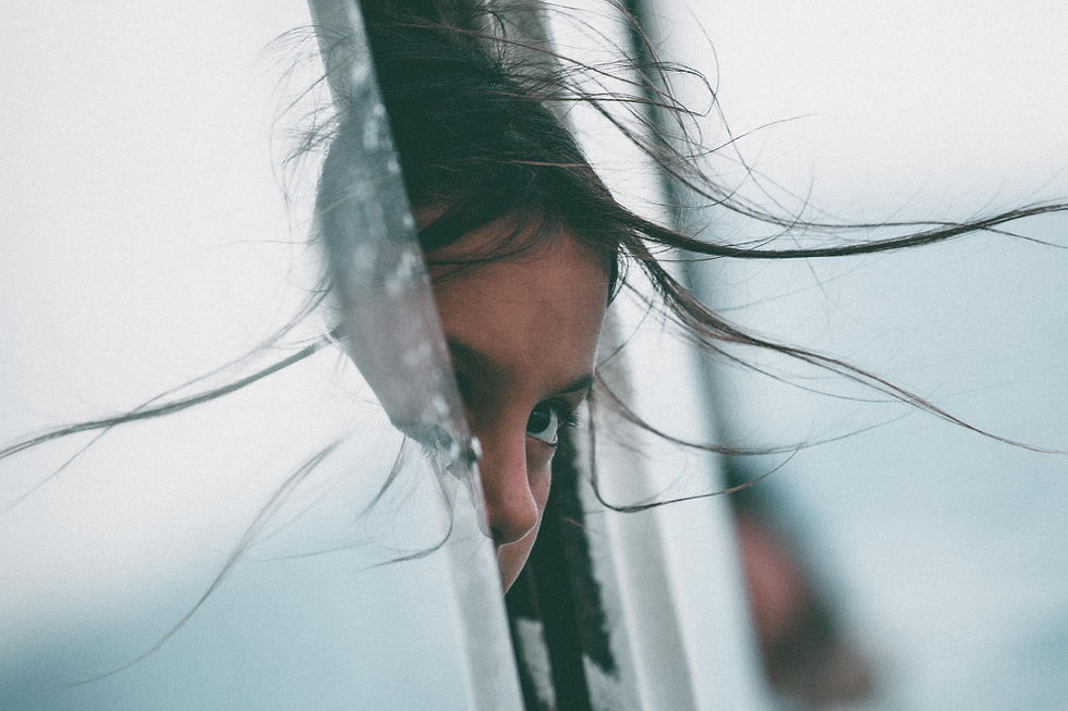 A shy young girl with windblown dark hair looks out from a glass door.