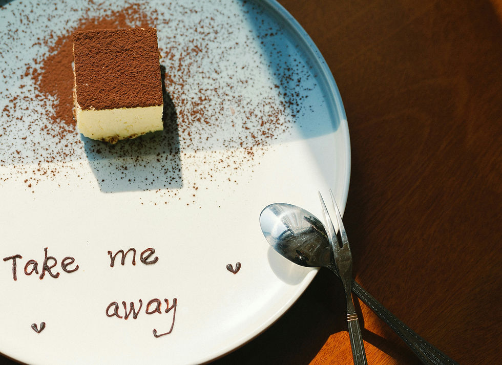 Photograph of a white, chocolate dusted dessert on a. round white pate with a small lip. "Take me away" is written in chocolate sauces with two hearts bordering. A spoon and two pronged fork are leaned against the plate. A cherry wood table is in background.