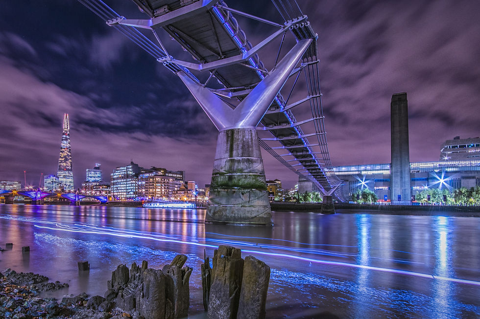 Night view of illuminated Millennium Bridge over the Thames, with London skyline, purple clouds, and blue light trails on water.