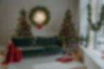 Festive room with two decorated Christmas trees, a "Happy New Year" wreath, green sofa with lights, red throw, gifts, and a snowy window view.