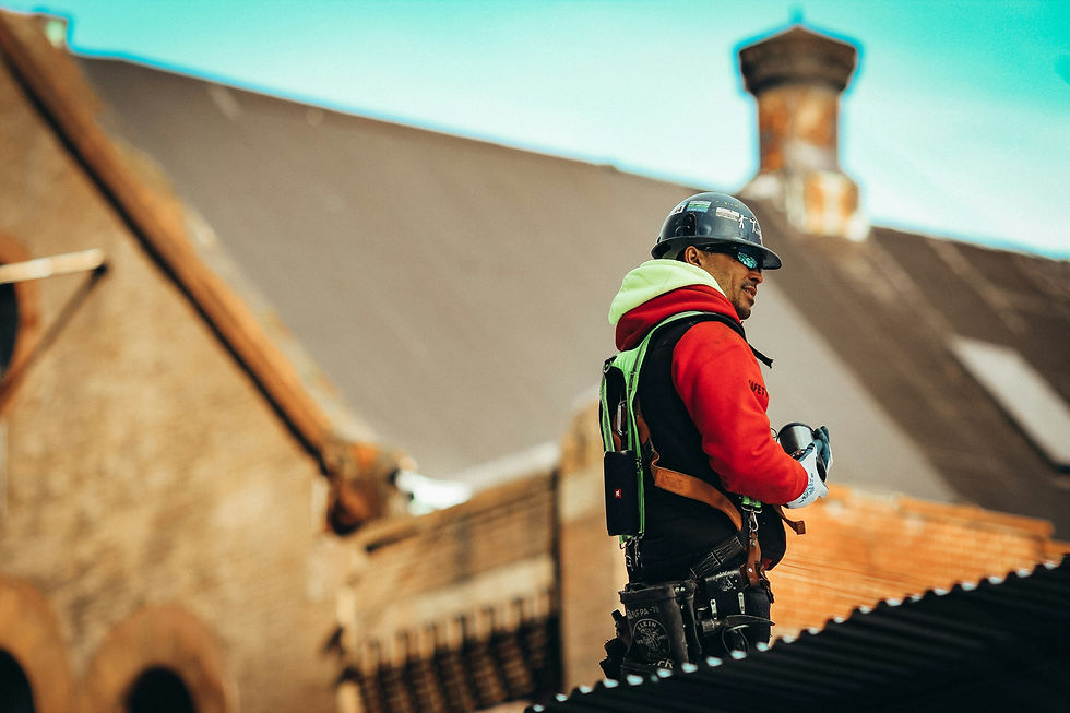 Construction worker in a red hoodie and safety gear stands on a roof, holding tools. Background shows an industrial building and blue sky.