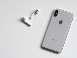 Grey iPhone and white AirPods on a white surface. The iPhone has Apple and "iPhone" logos visible; AirPods are placed near it. Minimalist setting.
