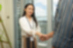 A woman wearing a white blouse and ID badge smiles and shakes hands with a person in a striped shirt, standing by a window with a city view.