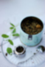 Tin of loose herbal tea labeled Lung Loving Brew with fresh mint leaves and a tea infuser on a clear plate