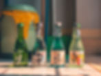 Four green and clear sake bottles with Japanese labels sit on a sunlit tiled floor. A yellow trash bag is visible in the background.
