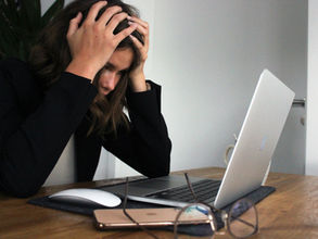 Person with hands on head, sitting at a wooden desk with a laptop, phone, and glasses. Appears frustrated. Background is plain white.