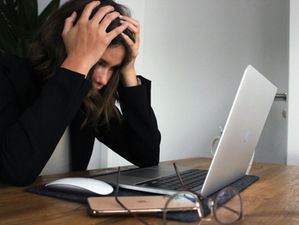 Person with hands on head, sitting at a wooden desk with a laptop, phone, and glasses. Appears frustrated. Background is plain white.