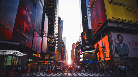 Times Square in New York City at sunset, with illuminated billboards, busy streets, and towering skyscrapers lining the avenue.