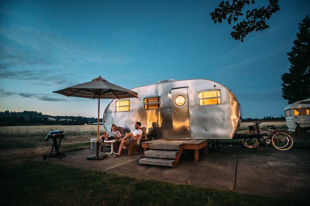 Young couple sit under umbrella outside Jetstream trailer hookup on cement pad in field campsite during late evening