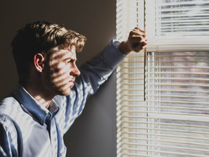 Man in blue shirt gazes out window, sunlight creating striped shadows on his face. The room is dim, mood appears contemplative.