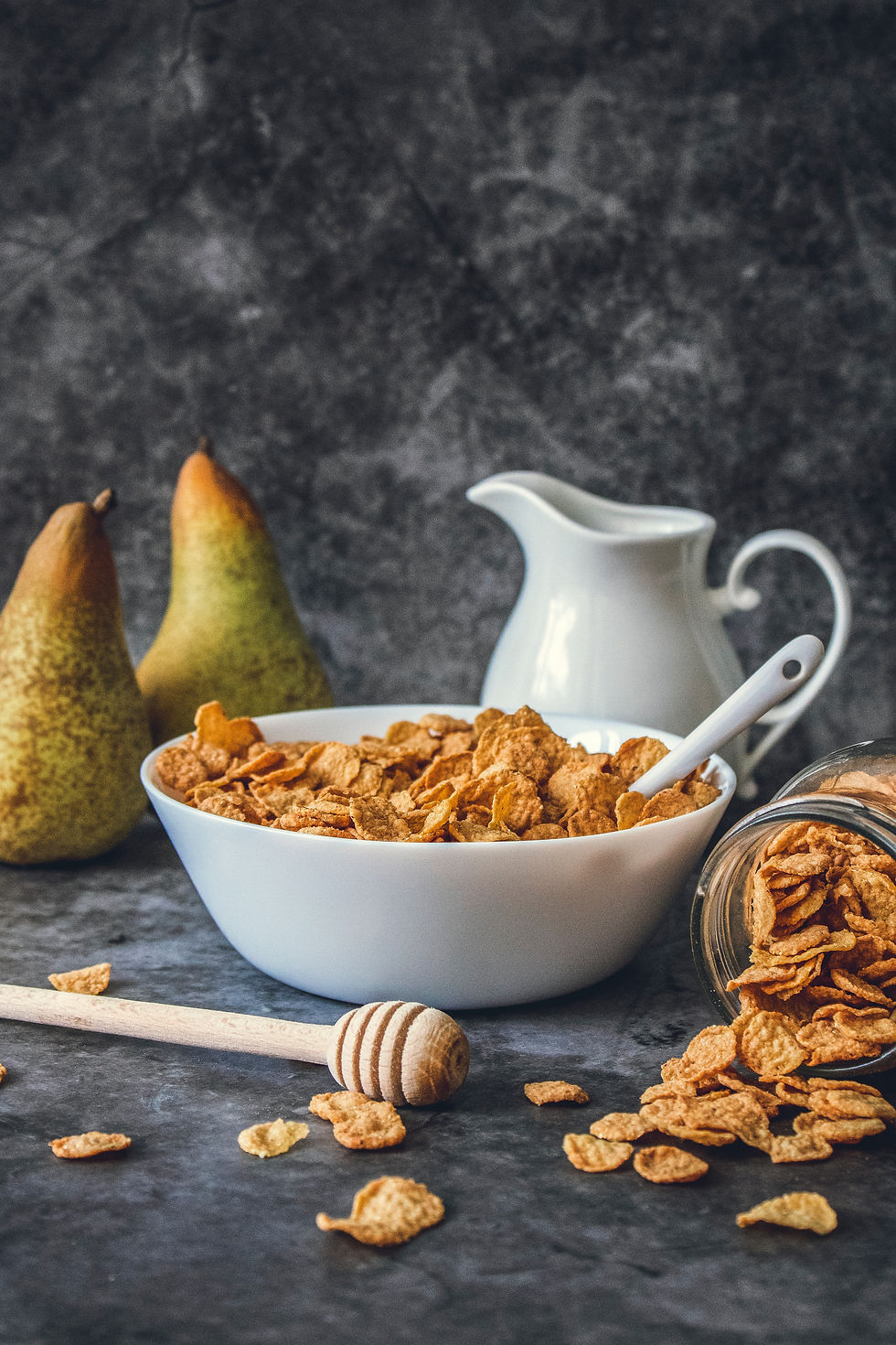 Wooden honey and syrup dipper stick on a slate gray backdrop with a white milk pitcher, two ripe pears, white cereal bowl with white spoon and crispy cereal bran flakes, and glass mason jar tipped on it's side with cereal flowing out and onto the table.