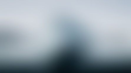 Massive white iceberg floats in calm dark Arctic water, with smaller ice chunks and hazy mountains under a pale sky.