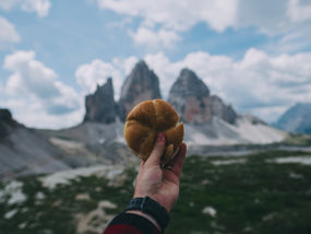 Hand holding a bread roll in front of mountain peaks under a cloudy blue sky. Scenic outdoor setting with a sense of adventure.