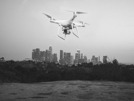 Drone hovers in the foreground with a city skyline in the background, set against a clear sky. Monochrome tones evoke a calm mood.